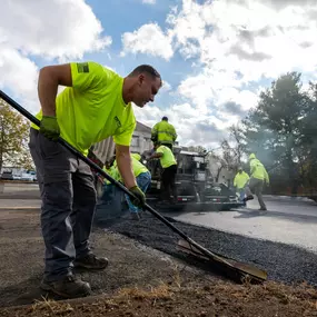 A worker smooths out the edge of newly laid asphalt while the rest of the team paves the road.