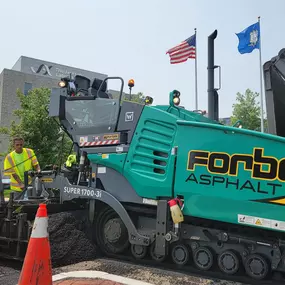 Forbes Asphalt team paving at a commercial site, with American and state flags flying in background.