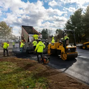 Full crew working together to pave a road, operating trucks, rollers, and compactors.