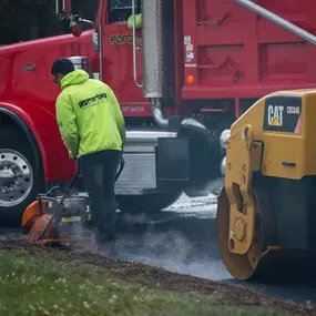 A Forbes Asphalt crew member operates a plate compactor near a red dump truck and CAT roller.