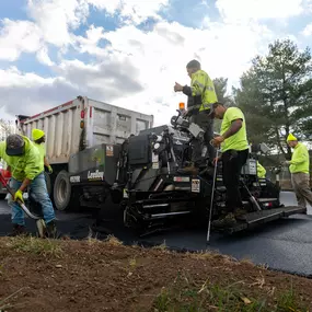 A crew works together on fresh asphalt, operating a LeeBoy paver beside a dump truck