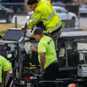 Two crew members operate the control panel of a paving machine during asphalt work
