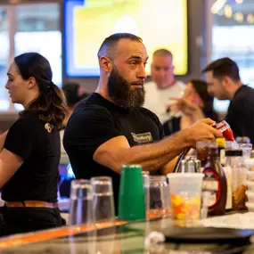A Brewbury bartender prepares drinks behind the bar with focus and flair, framed by cocktail ingredients and a busy team during service.