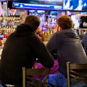 Two guests relax at Brewbury’s bar, facing a wall of taps and TVs while enjoying drinks and good conversation in a casual setting.