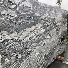 A large slab of natural stone, likely granite or marble, stands upright, showcasing its dramatic grey, black, and white wavy patterns. Its polished surface reflects subtle light.