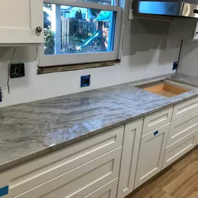 A kitchen under renovation features new grey natural stone countertops with prominent veining, installed over white base cabinets. A sink cutout and exposed electrical outlets indicate ongoing work.
