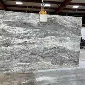 A large, stunning slab of natural stone, featuring dynamic grey, white, and brown swirling patterns, is carefully hoisted by machinery within a warehouse, ready for fabrication.