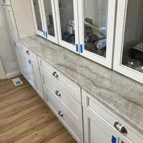 A long run of newly installed light-colored natural stone countertops with subtle brown and grey veining, complementing white shaker-style base cabinets and glass-front upper cabinets.