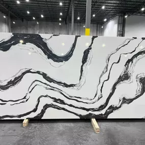 A striking large slab of white stone, likely quartz, features bold, dramatic black and grey swirling veins, resembling natural marble, showcased in a warehouse setting.