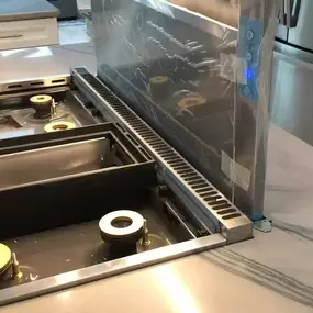 A close-up view of a high-end kitchen island featuring a modern cooktop with brass burners and an integrated downdraft ventilation system, set into a light-colored, veined stone countertop.