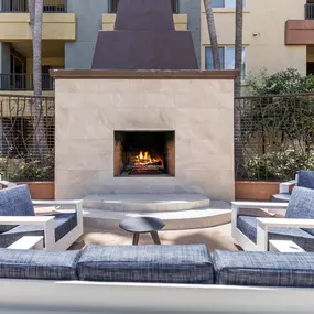 A fireplace is in the middle of a patio with blue cushioned chairs around it.