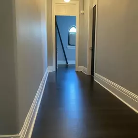 This freshly renovated hallway showcases Arber Construction Group’s signature attention to detail. Featuring rich, dark wood-style flooring, crisp white baseboards, and smooth, modern gray walls, the space is elevated by clean lines and a timeless pendant light fixture. Perfectly framed by high ceilings and a glimpse of the arched window beyond, this project reflects our commitment to craftsmanship and sophisticated design in every corner.