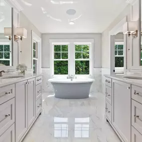 A spacious, bright master bathroom features dual white vanities with marble countertops and mirrors, facing a light gray freestanding tub positioned beneath a large window offering green outdoor views.
