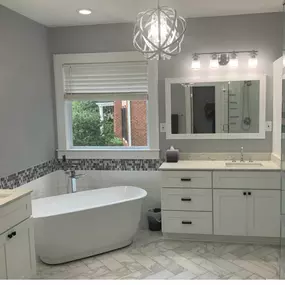 A modern bathroom features a freestanding tub, double vanity with white cabinets, and light gray walls. Herringbone tile flooring and a mosaic backsplash add texture.