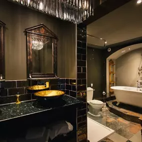 This opulent bathroom showcases dark walls and black subway tiles, complemented by ornate mirrors and striking gold basin sinks. A freestanding white tub occupies an arched alcove, enhancing the luxurious ambiance.