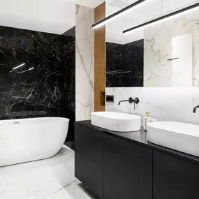 This modern bathroom features a dramatic black marble accent wall behind a white freestanding tub. Dual white vessel sinks sit atop a sleek black vanity, contrasted by white marble walls and flooring.