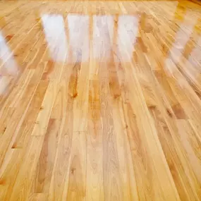 This image showcases a beautifully polished hardwood floor with a warm, natural wood grain and a glossy finish. Reflections from overhead lighting create bright streaks across the smooth surface.