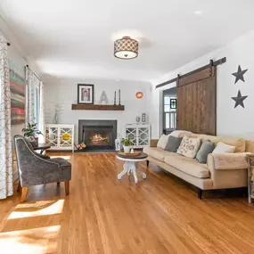 A cozy living room features warm honey oak floors, a fireplace, and a beige sofa. The room is bright with natural light, showcasing a rustic barn door and white walls.