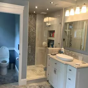 A modern bathroom features a white vanity with an oval sink, a glass shower with mosaic tile accents, and light-colored large format tiles. A separate toilet room is visible through an open doorway.