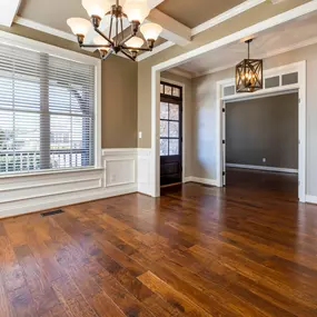 An empty living room features rich, dark parquet flooring, reflecting light from a window. The room is partially visible, showing a doorway and a glimpse into adjacent spaces.