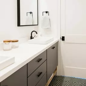 A modern bathroom features a dark gray vanity with a white countertop and two rectangular vessel sinks. Black fixtures and pulls complement the clean aesthetic, with a geometric pattern on the floor.