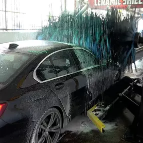A close-up of the wash tunnel in action at Speedwash Catalina. The image shows a black BMW receiving a high-pressure foam wash with spinning brushes and overhead sprayers, demonstrating premium auto wash equipment and service quality.