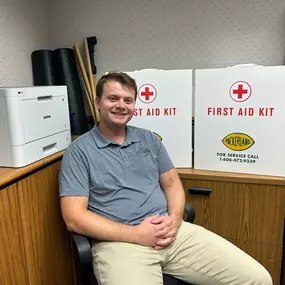 A Packerland employee wearing a grey collared shirt and khaki pants sits on an office chair with his hands casually folded on his lap. Behind him are two white first aid cabinets with the Packerland logo, which is a yellow football with green text.