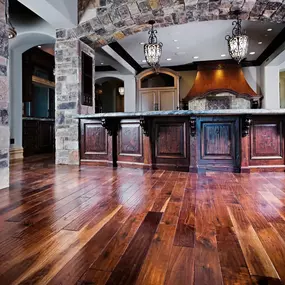 A luxurious kitchen with polished hardwood floors, a stone archway, a large dark wood island, and hanging bronze light fixtures.