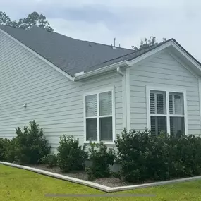 The side of a modern home with light green siding, a black roof, and a screened-in porch at the back, surrounded by well-maintained landscaping and bushes.