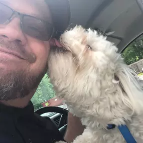 A close-up selfie of a man with a beard and sunglasses smiling while holding a small, fluffy white dog that is licking his cheek.