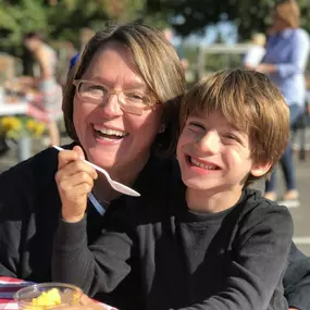 A happy-looking woman with glasses and her son are smiling at the camera for a close-up photo. They are seated at a table with a red and white checkered tablecloth.