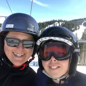 A woman with sunglasses and a son with ski goggles are smiling at the camera for a selfie. They are on a ski lift, wearing helmets, with snow-covered mountains in the background.