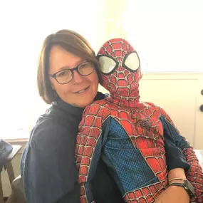 A professional-looking woman with glasses and brown hair is embracing a child in a Spider-Man costume, who has his back to the camera. They are both smiling and hugging.