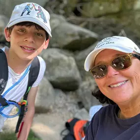 A selfie of a smiling woman with sunglasses and a son with a Star Wars hat. They are wearing casual clothes and backpacks, posing outdoors with large rocks in the background.