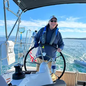 A professional-looking woman with a white hat and blue sweatshirt is smiling while standing at the helm of a sailboat. She is wearing a life vest, with the open water in the background.