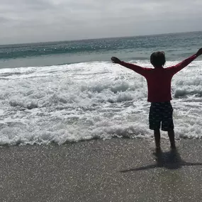A child stands on a sandy beach with arms outstretched, facing the ocean. The silhouette is in front of white-capped waves, with the horizon and a cloudy sky in the background.
