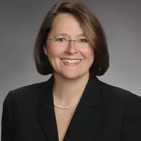A professional headshot of a smiling middle-aged woman with short brown hair, glasses, and a pearl necklace. She is wearing a black blazer in front of a gray studio background.