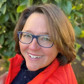 A professional headshot of a smiling woman with stylish blue glasses and short brown hair. She is wearing a red vest over a dark blue shirt with green foliage in the background.
