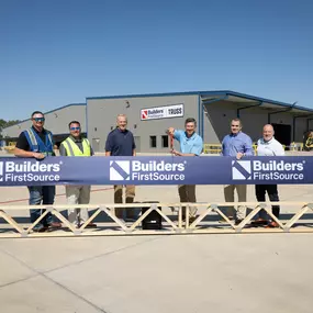 Six Builders FirstSource team members stand in front of a large banner displaying the company logo at the Conroe, TX manufacturing facility. They are spaced evenly behind a wooden truss, holding a ribbon-cutting tool as they celebrate the opening. The 