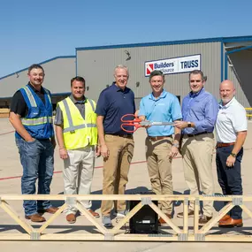 A group of six Builders FirstSource team members stands together for a ribbon-cutting ceremony outside the new Conroe, TX manufacturing facility. The group holds a large pair of ceremonial scissors in front of a truss framework, with a Builders FirstSource 