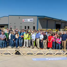 A large group of Builders FirstSource team members and affiliates gather outdoors in front of a newly constructed building labeled 