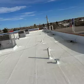 A wide shot of a flat, white coated roof on a commercial building, with a Marathon gas station visible in the background. The roof is clean and uniform, with various pipe vents and a single HVAC unit.