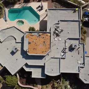 An aerial view of a large residential property with a multi-section flat roof undergoing a major replacement. The image shows a central section of the roof where the old material has been completely removed, revealing the orange-brown base layer.