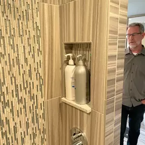 This image shows a newly renovated bathroom. The walk-in shower features tiled walls and a river stone floor. There is also a double vanity with an illuminated mirror on a dark green accent wall.