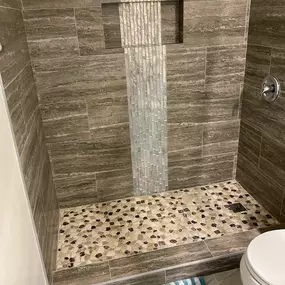 This image shows a newly renovated walk-in shower with a custom-tiled niche for bath products. The walls are tiled with a natural wood-grain pattern and a vertical strip of smaller mosaic tiles. The shower floor is a stone pebble mosaic.