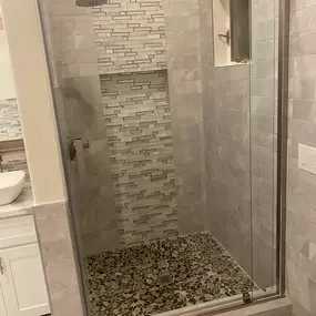 This image shows a newly renovated bathroom walk-in shower. The walls are finished with a striped pattern of brown tiles, a vertical mosaic of small gray tiles, and a custom river stone floor. A toilet is partially visible on the right.