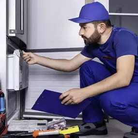 A technician in a blue uniform examining the inside of a refrigerator with tools and clipboard in hand, representing professional appliance diagnostics