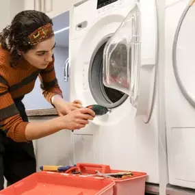 A woman using a power drill to work on the door of a front load washing machine, with a red toolbox in front