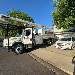 A white Clark's Tree Works bucket truck is parked on a suburban driveway, with a white sedan parked next to it. The truck, featuring the company logo and contact details, is ready for tree service operations.