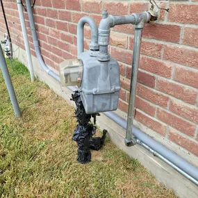 This is a close-up, outdoor photo of a gas meter mounted on a red brick wall. The meter has been damaged, with the base of the pipework appearing to be severely burned and melted.